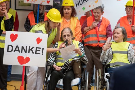 Menschen mit Warnwesten, Bauhelmen und Rollstühlen sitzen bzw. stehen. Ein Schild mit der Aufschrift "Danke" ist zu sehen.