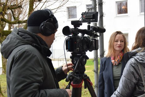  Eine Frau mit buntem Halstuch sieht eine Frau mit einer silber-färbigen Jacke an. Ein Mann mit Haube, von dem man den Rücken sieht, filmt die Frau mit dem bunten Halstuch.