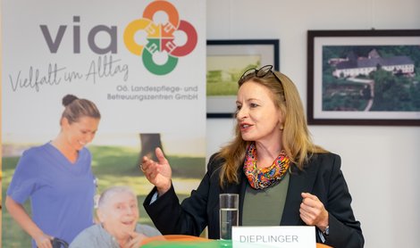 Eine Frau steht hinter einem Stehtisch und erhebt die Hände. Im Hintergrund ist das gelb-orange-rot-grüne Logo der LPBZ GmbH zu sehen.